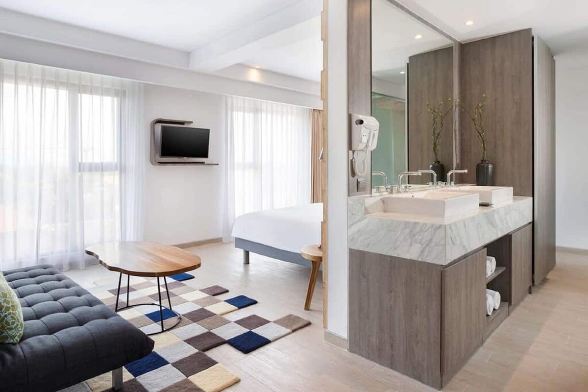 Ein modernes Hotelzimmer mit einem großen Bett, einer Sitzecke mit einer Couch und einem kleinen Tisch, einem Badezimmer mit einem großen Spiegel und einem Waschtisch sowie einem an der Wand montierten Fernseher.