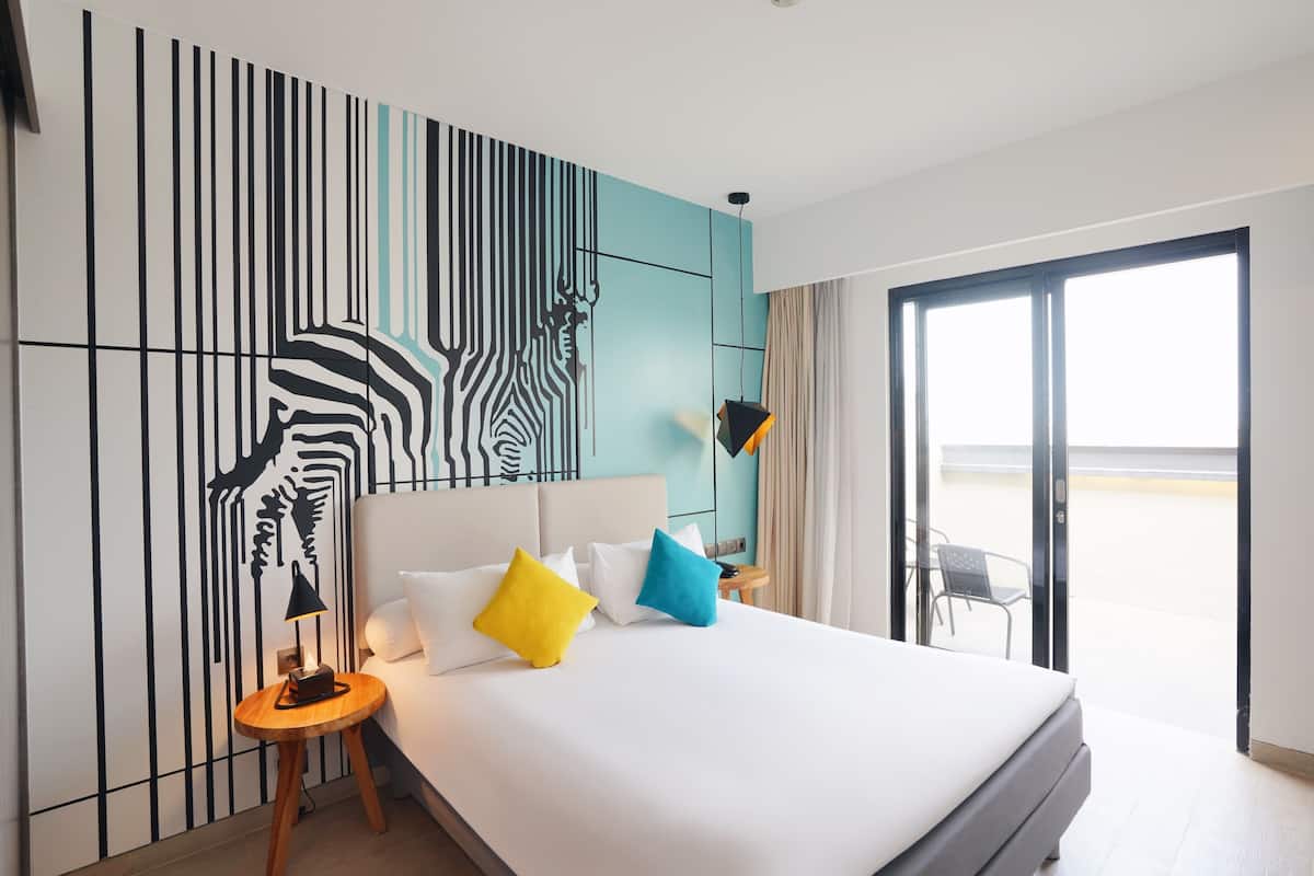Ein Hotelzimmer mit einem großen Bett, einem Zebra-Wandbild, einem Balkon mit einem Stuhl und Blick ins Freie.