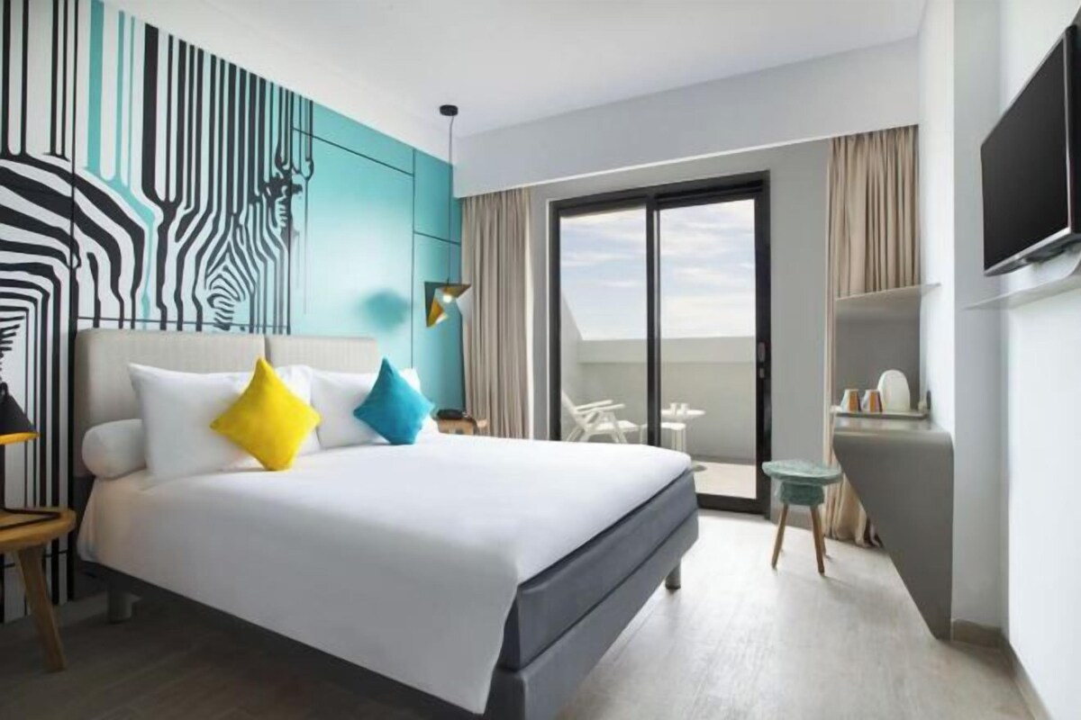 A modern hotel room with a large bed, a view to the outside, a small table, and a TV mounted on the wall.