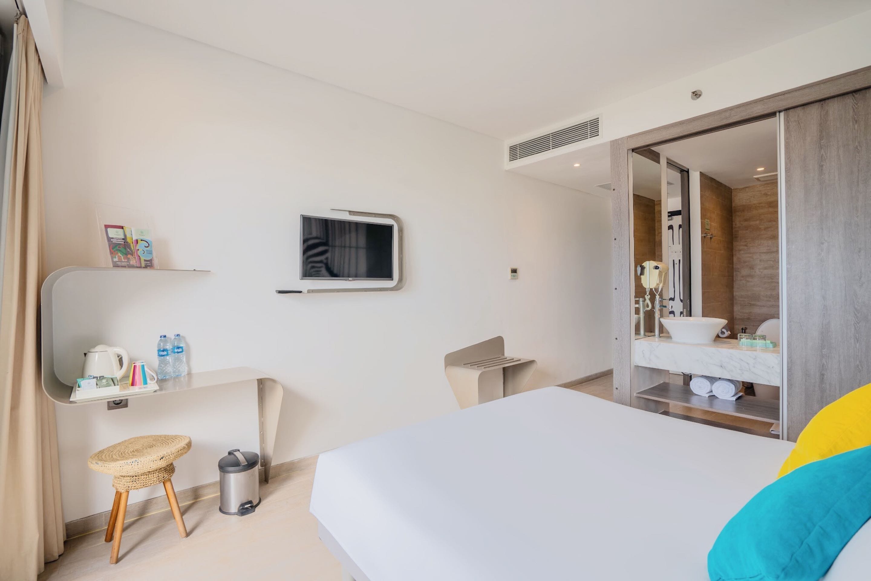 A hotel room with a bed, a TV, a small table with a kettle and water bottles, a trash bin, a wicker stool, and a bathroom with a mirror and sink.