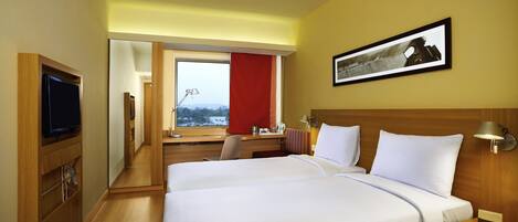 A hotel room with two beds, a TV, a desk, and a window with a red curtain.
