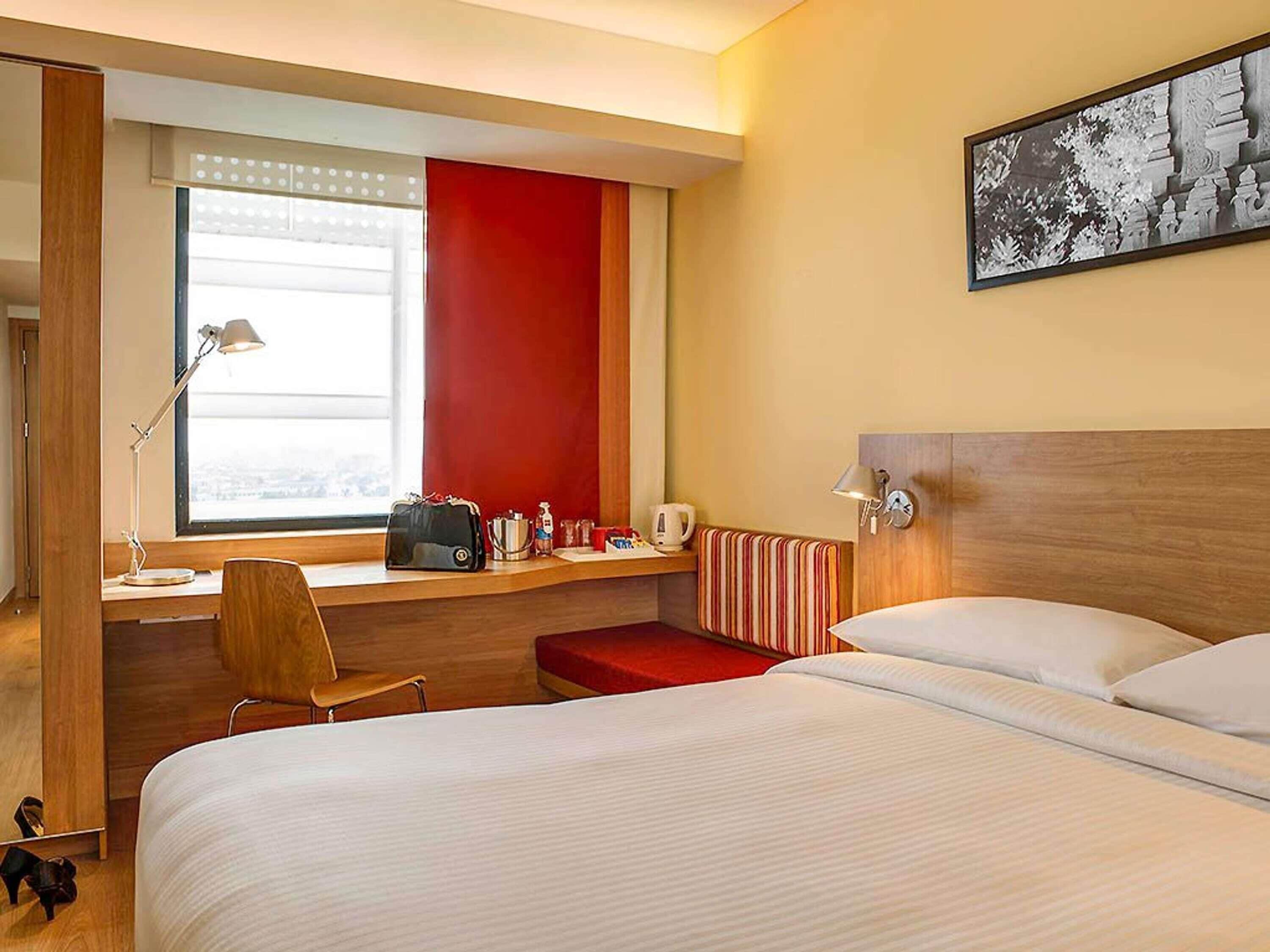 Une chambre d’hôtel avec un lit, un bureau et une chaise, une fenêtre et un tableau au mur.