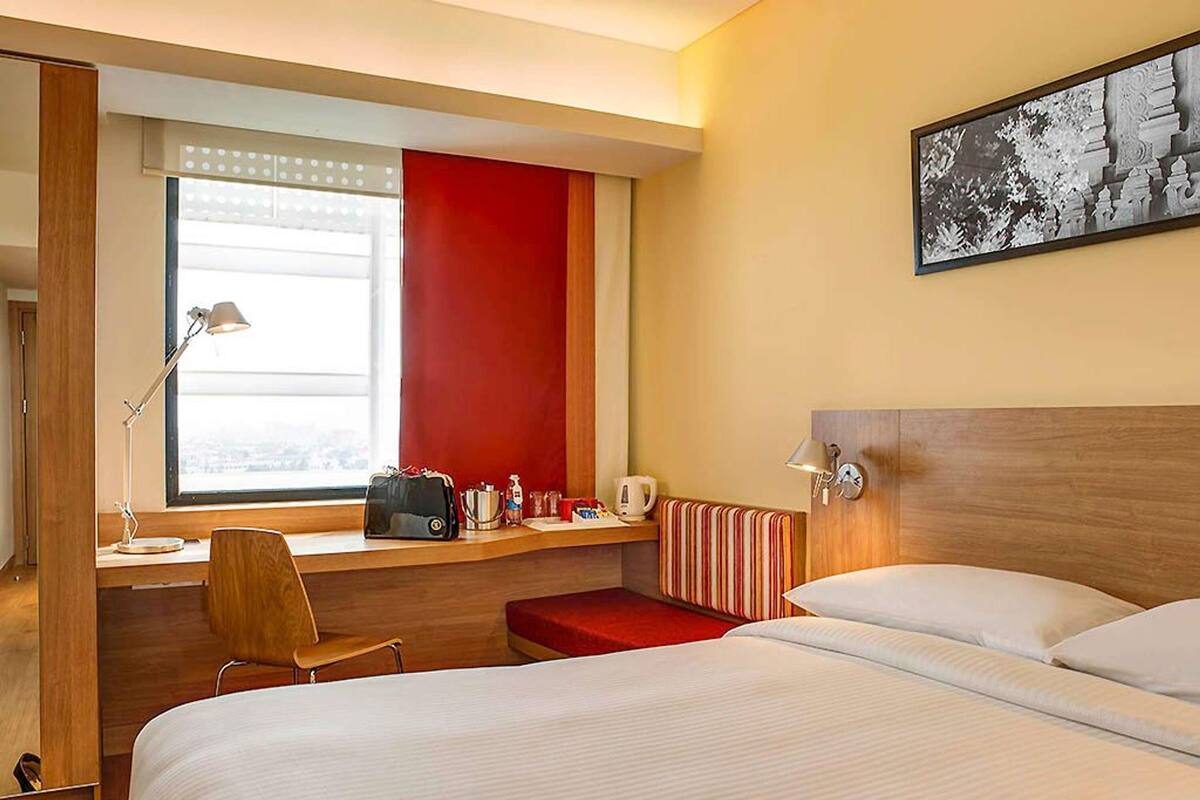 A hotel room with a bed, a desk with a chair, a window, and a painting on the wall.