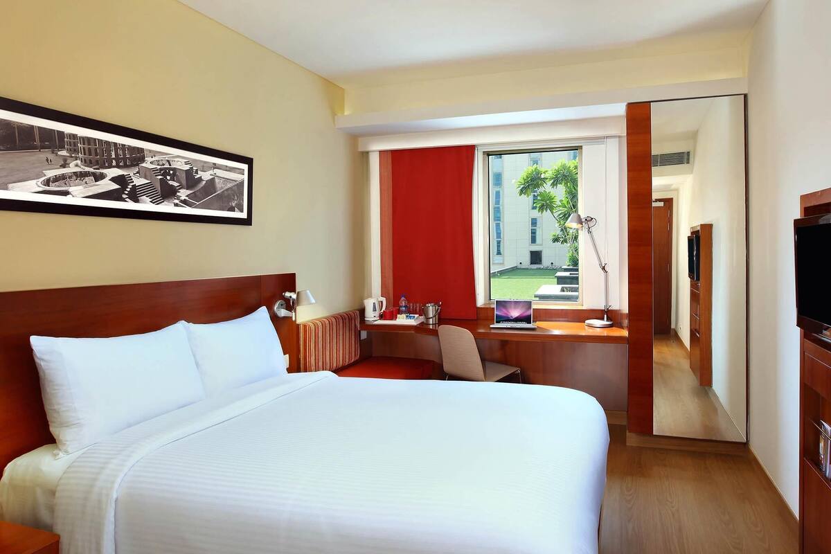 A hotel room with a large bed, a desk with a laptop, a TV, and a window with a red curtain.