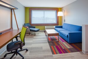 A modern hotel room with a large window, a blue sofa, a desk with a chair, and a colorful striped rug. - Holiday Inn Express & Suites Des Moines Downtown by IHG (Des Moines)