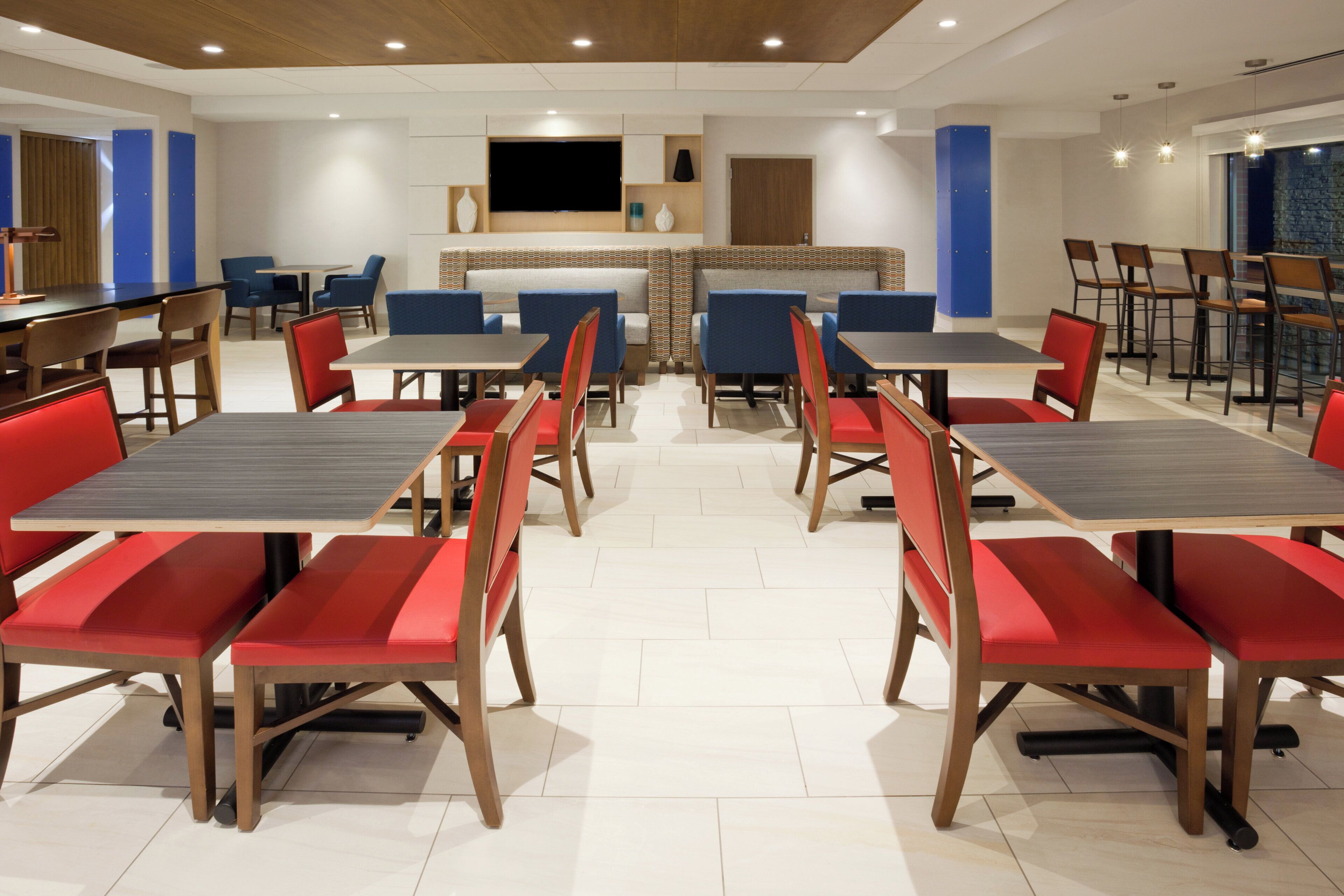 A modern dining area with multiple tables and chairs, a flat-screen TV mounted on the wall, and a patterned wall divider.