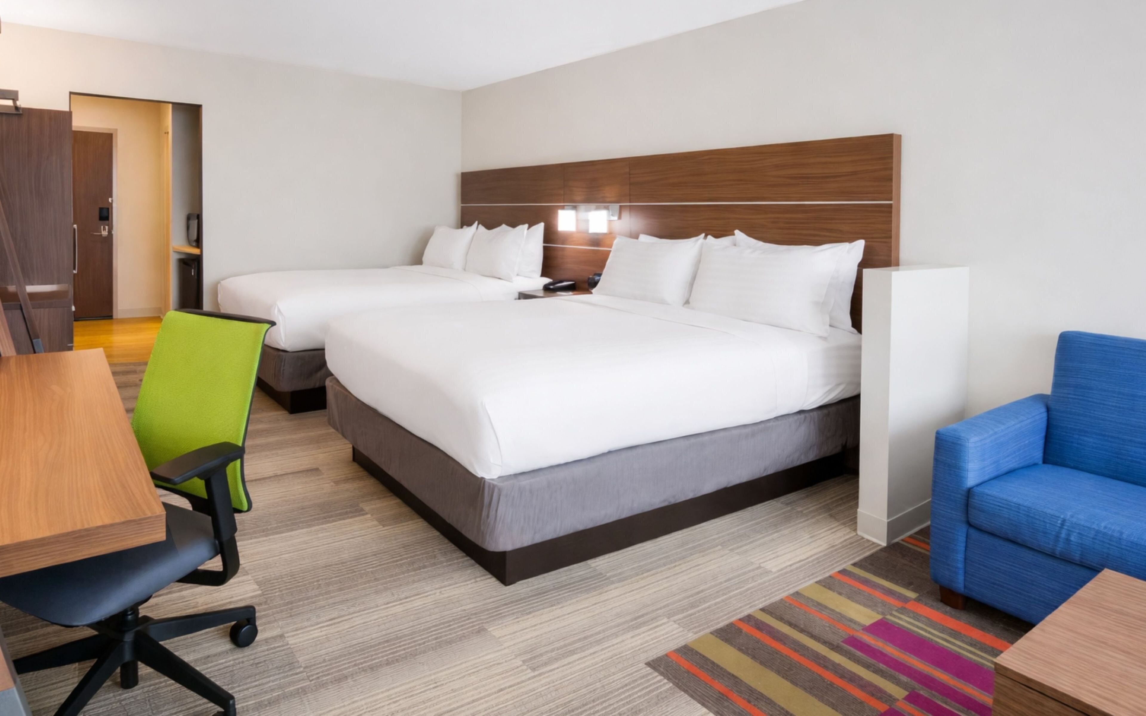 A hotel room with two beds, a desk, a chair, a sofa, and a colorful rug.