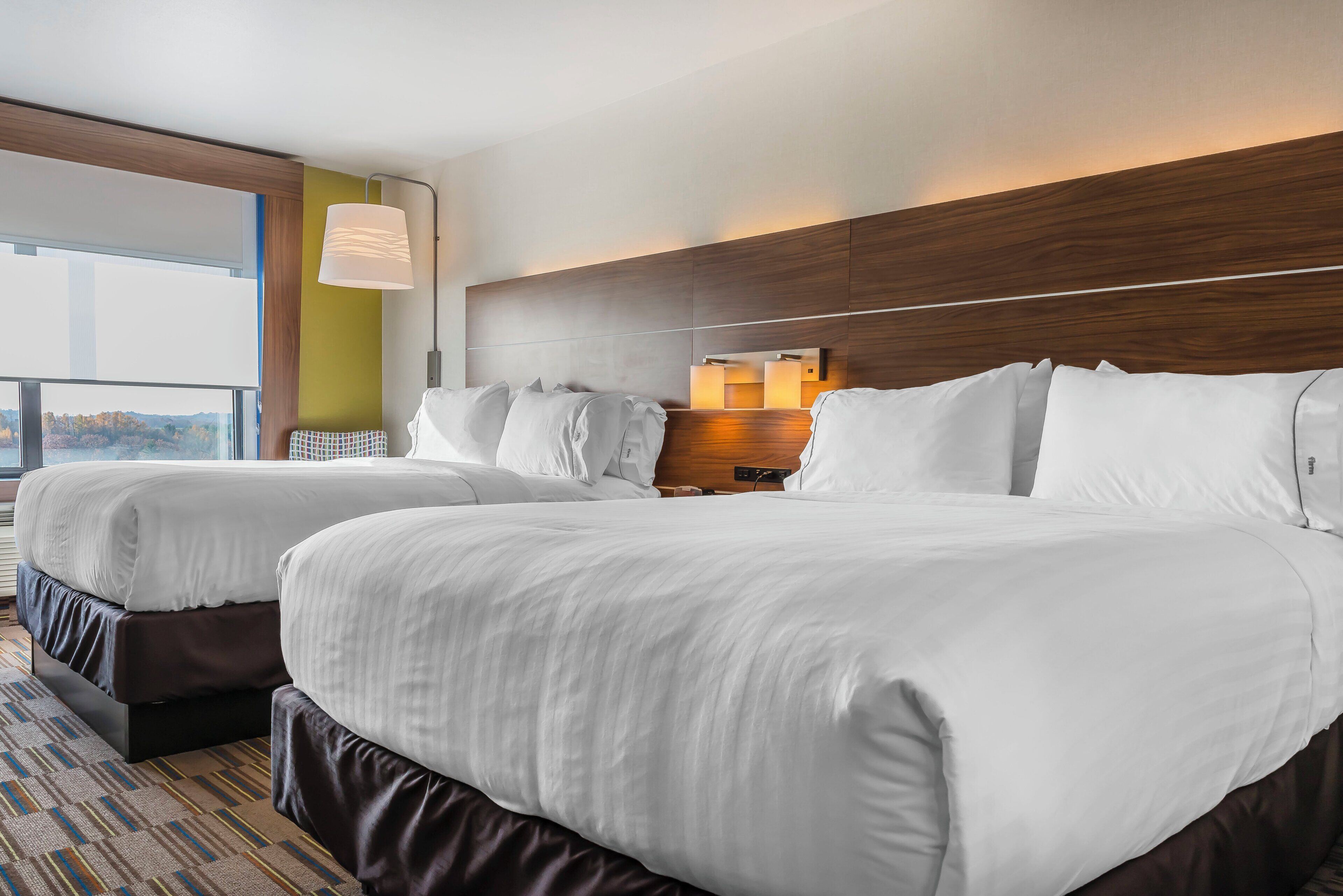 Habitación de hotel con dos camas, un ventanal grande, cabecero de madera y una mesita de noche.
