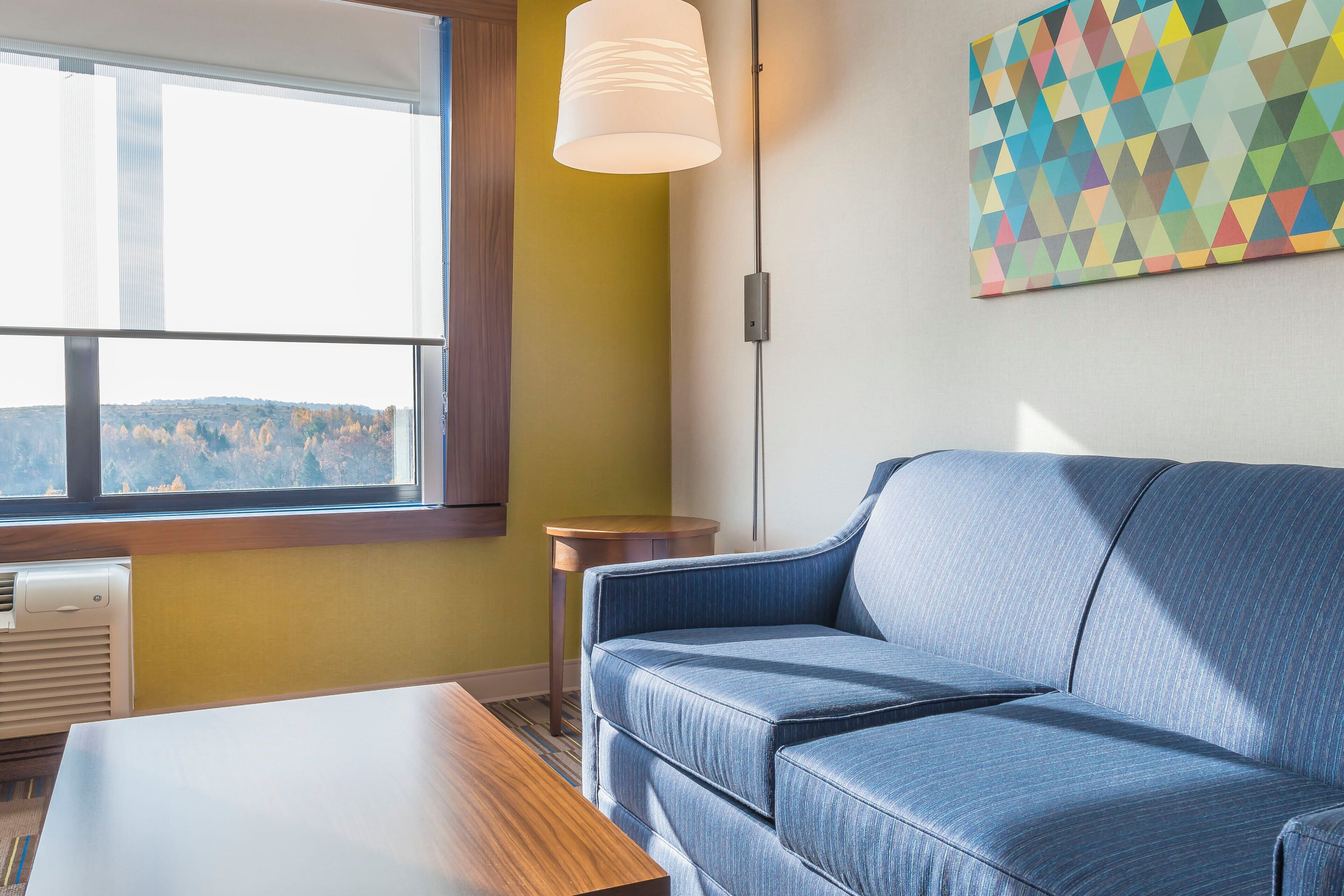 Une pièce avec un canapé bleu, une table en bois, une fenêtre avec des stores, une lampe fixée au mur et un tableau aux motifs géométriques.
