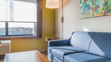 Une pièce avec un canapé bleu, une table en bois, une fenêtre avec des stores, une lampe fixée au mur et un tableau aux motifs géométriques.