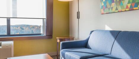 A room with a blue sofa, a wooden table, a window with blinds, a wall-mounted lamp, and a geometric painting.