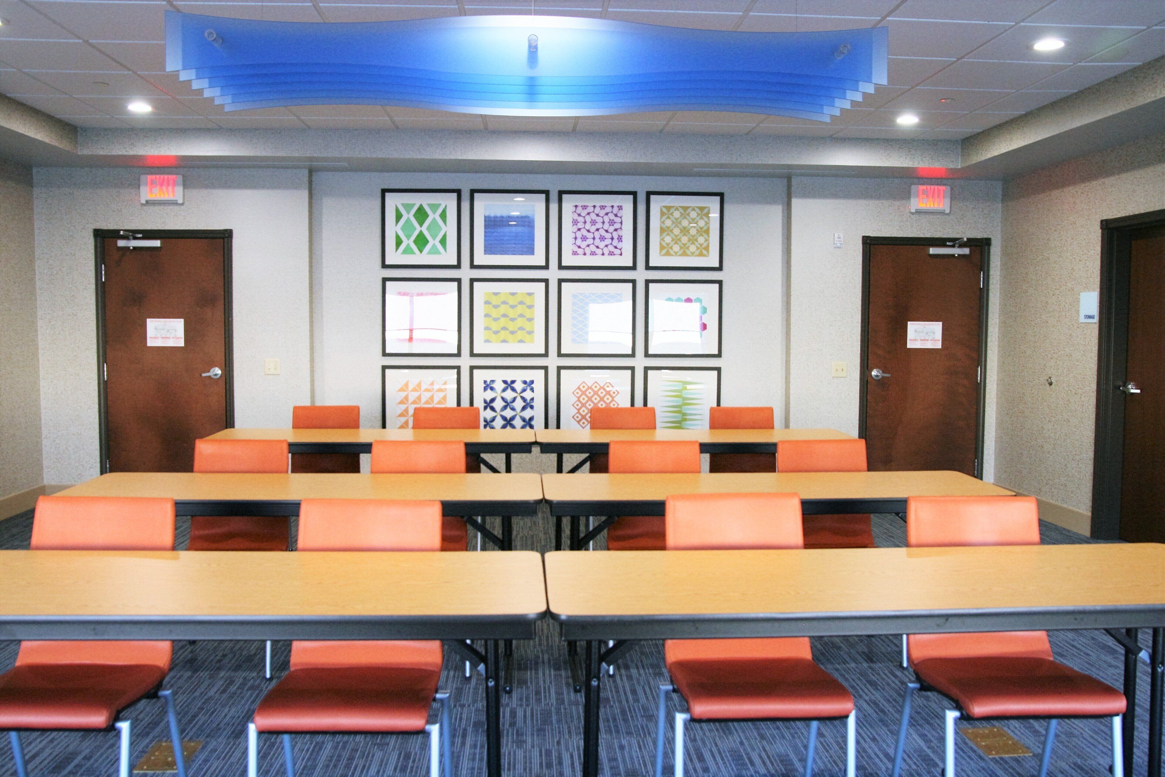 A conference room with multiple tables and chairs, framed artwork on the wall, and exit signs.