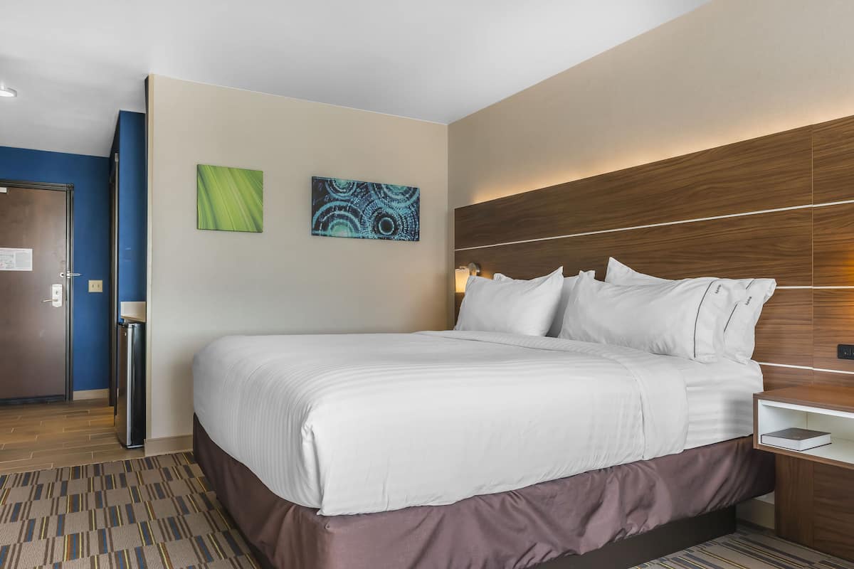 A hotel room with a large bed, a wooden headboard, two wall art pieces, a nightstand, and a door to the left.