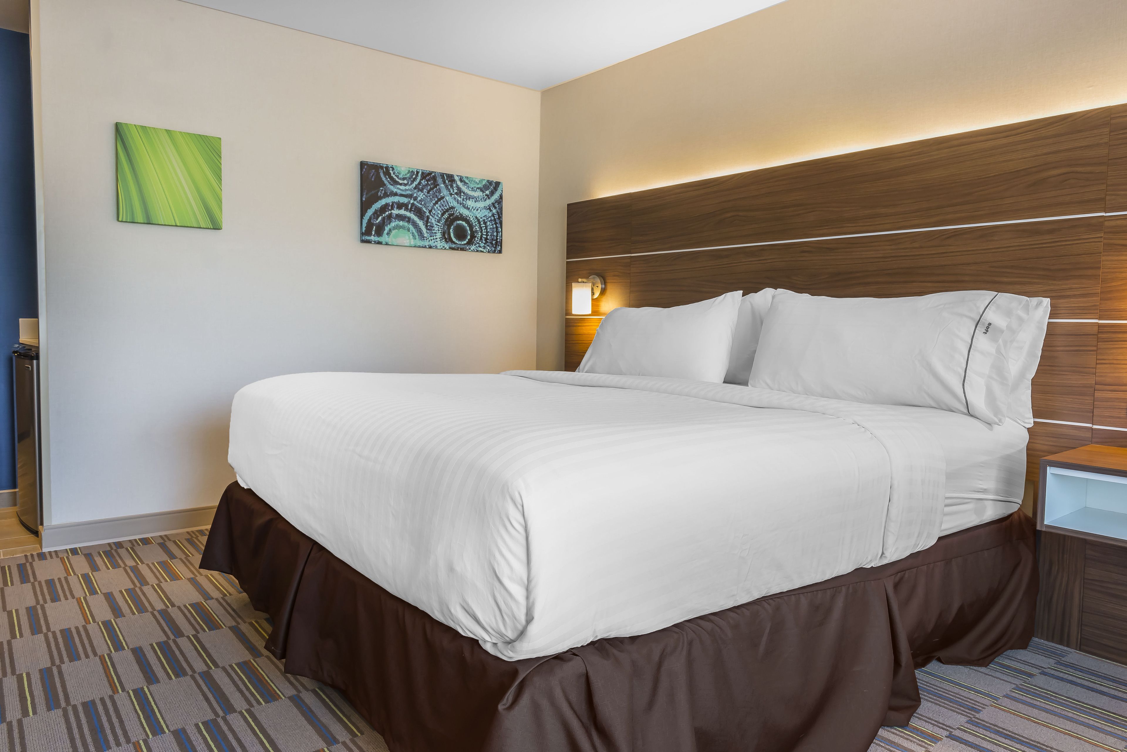 A hotel room with a large bed, a wooden headboard, two wall-mounted artworks, and a nightstand with a lamp.
