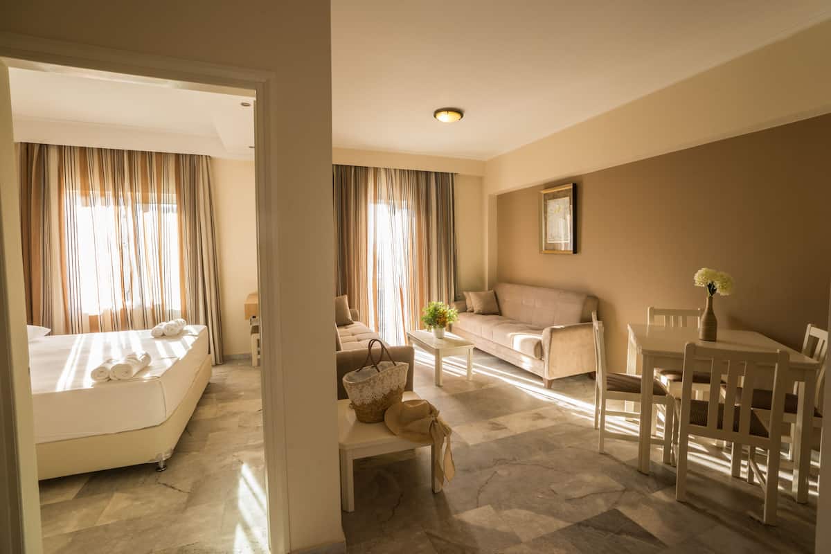 A hotel room with a bed, a sofa, a dining table, and a chair. There is a window with curtains, a lamp, and a vase with flowers on the table.