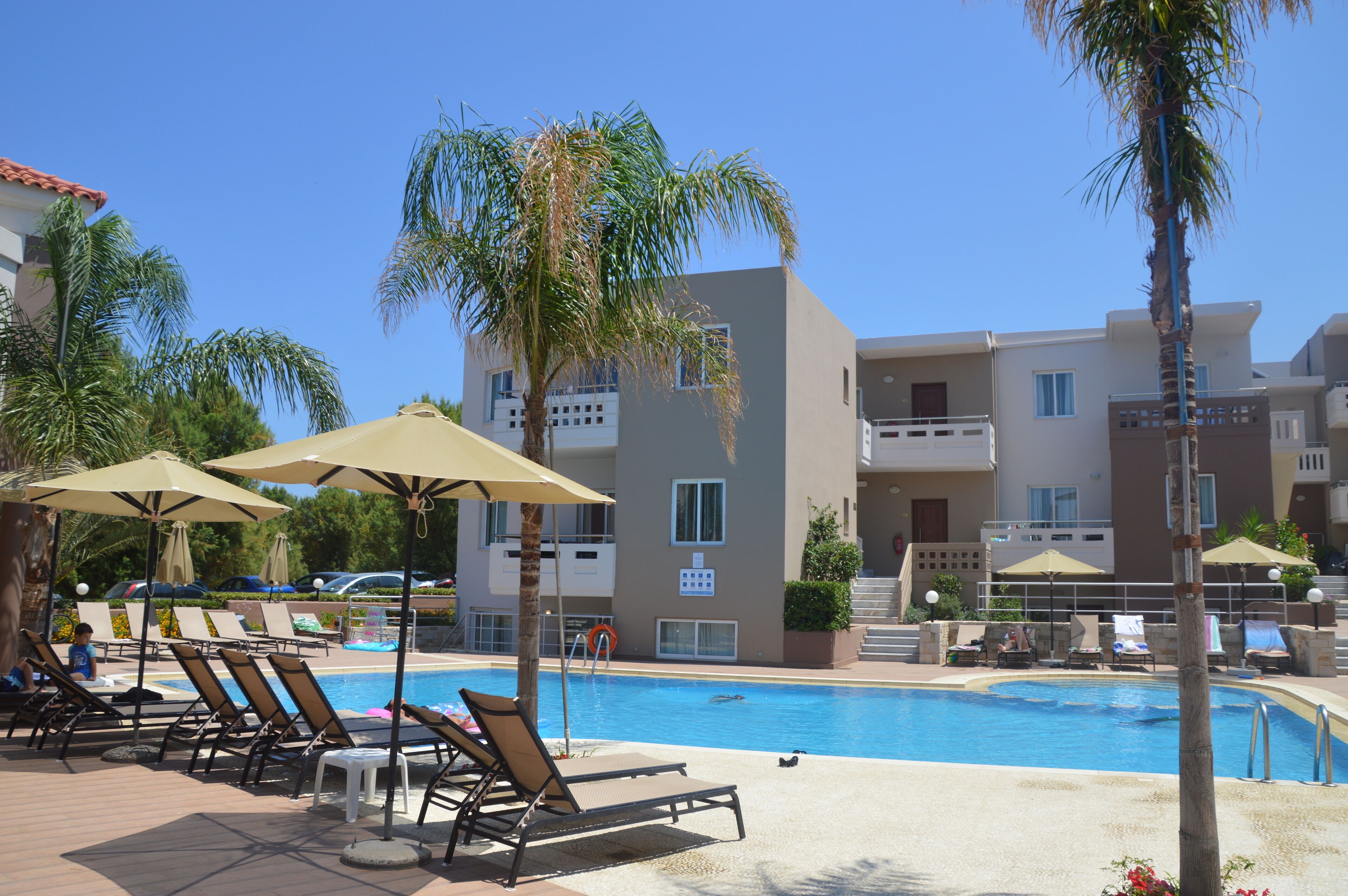 A pool area with lounge chairs and umbrellas, surrounded by palm trees and a building with balconies.