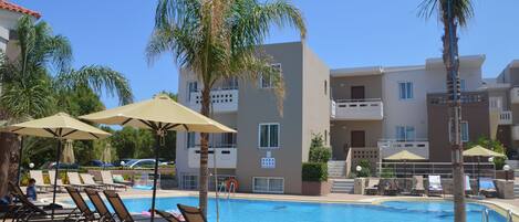 A pool area with lounge chairs and umbrellas, surrounded by palm trees and a building with balconies.