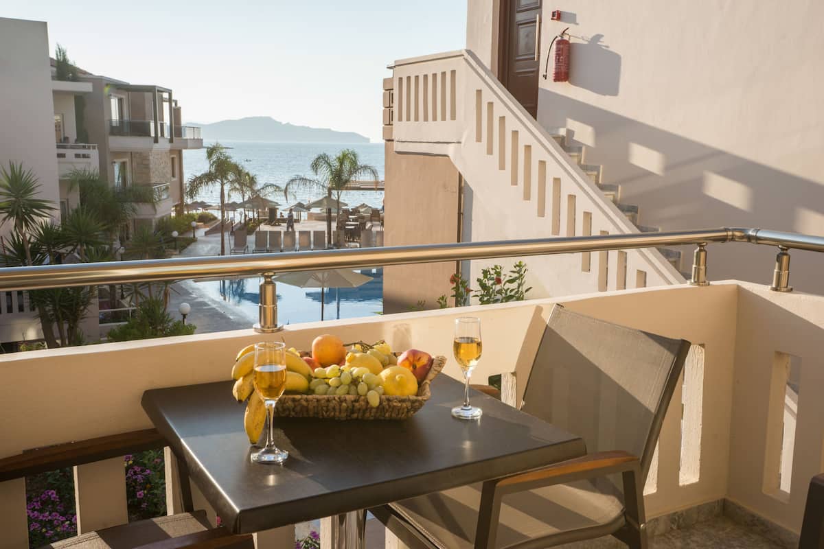A balcony with a table set for two, a fruit basket, and a glass of wine. The view includes a pool and a beach with umbrellas.