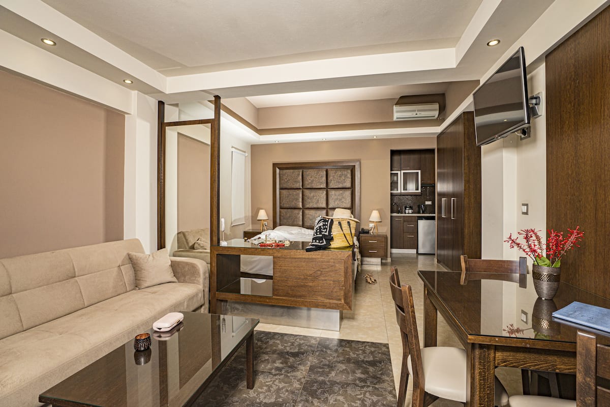 A modern hotel room with a sofa, a glass dining table, a wooden desk, a bed with a nightstand, a TV mounted on the wall, and a kitchen area in the background.