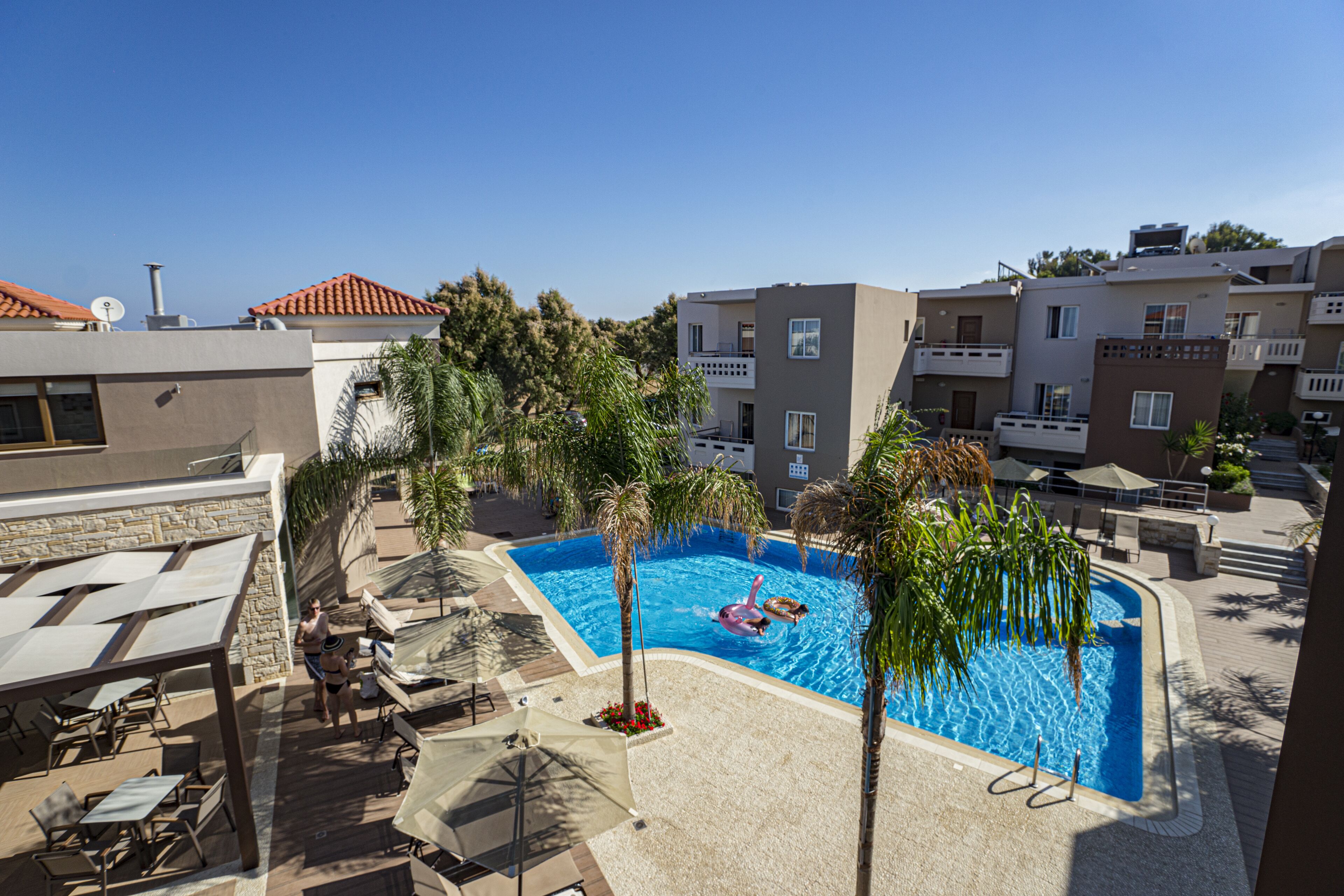 Zona de piscina con tumbonas y sombrillas, rodeada de edificios con balcones y un cielo despejado.