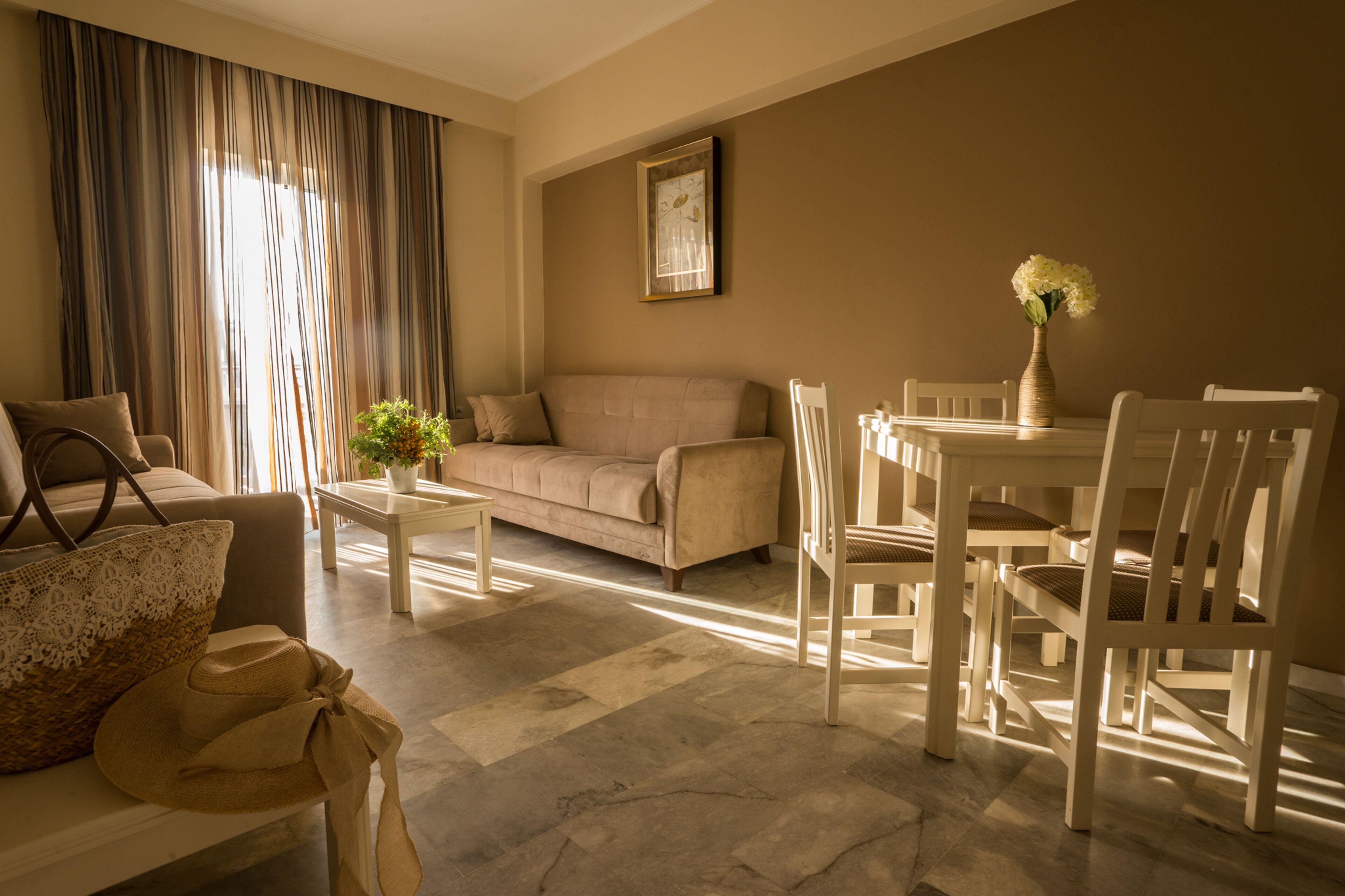 Un salon avec un canapé beige, une table à manger avec des chaises blanches, un vase rempli de fleurs et une grande fenêtre avec des rideaux.