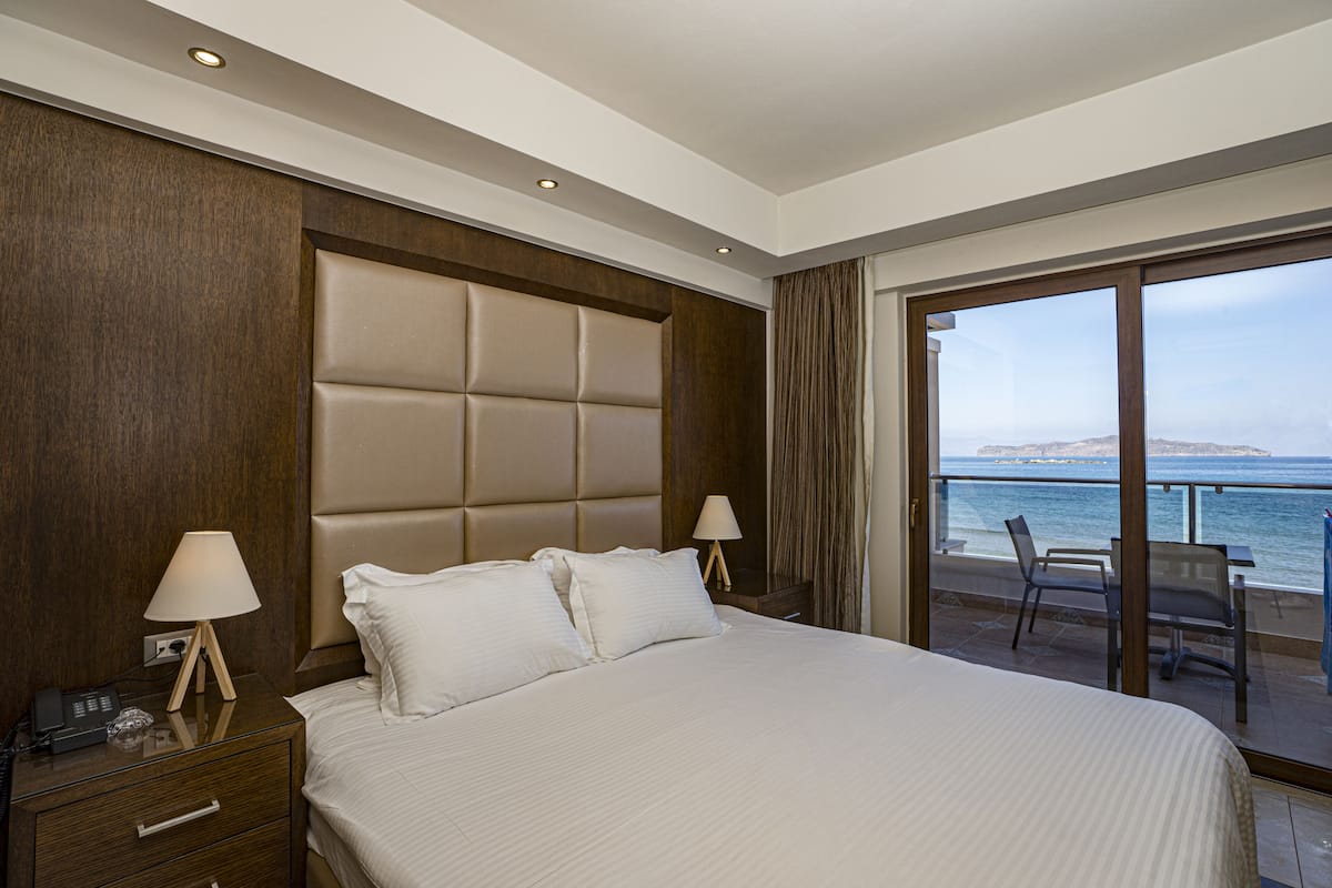 A hotel room with a large bed, two bedside tables with lamps, a wooden headboard, and a balcony with a view of the sea.