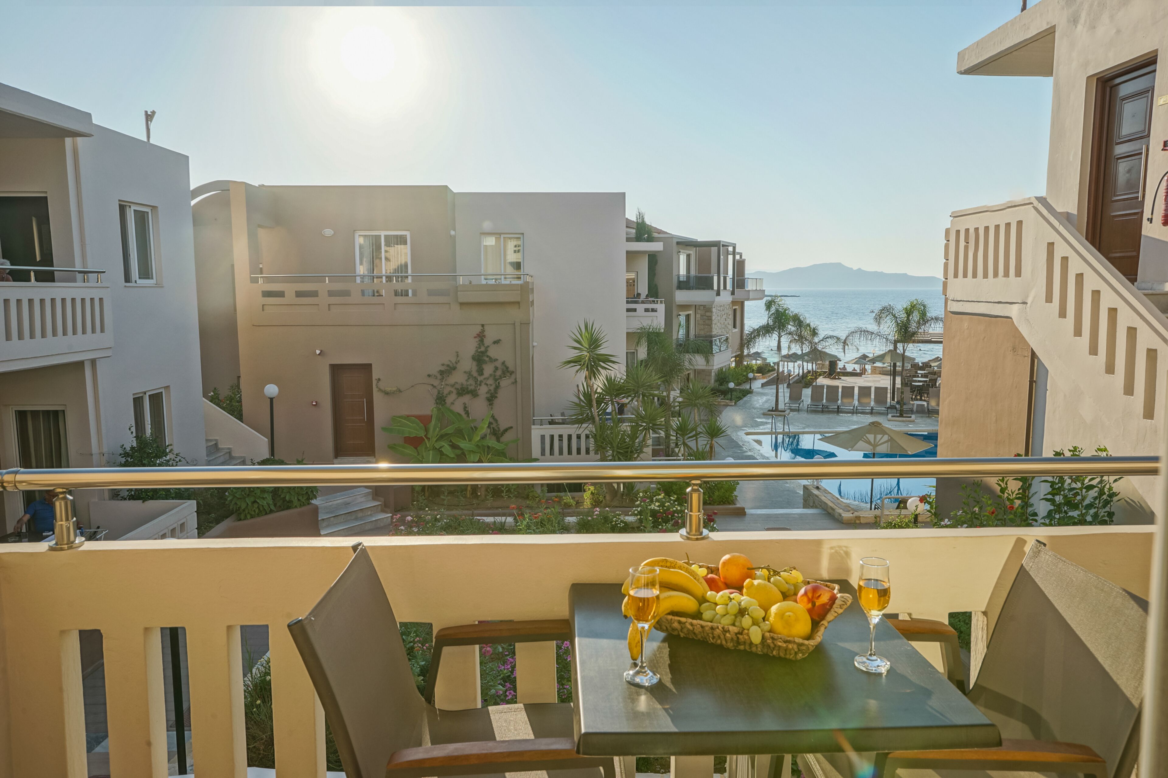 Un balcón con una mesa dispuesta con frutas y una copa de vino, con vistas a una piscina y una playa.