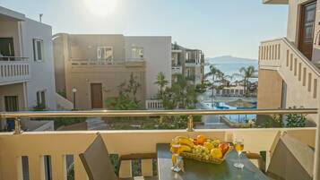 A balcony with a table set with fruits and a glass of wine, overlooking a pool and a beach.
