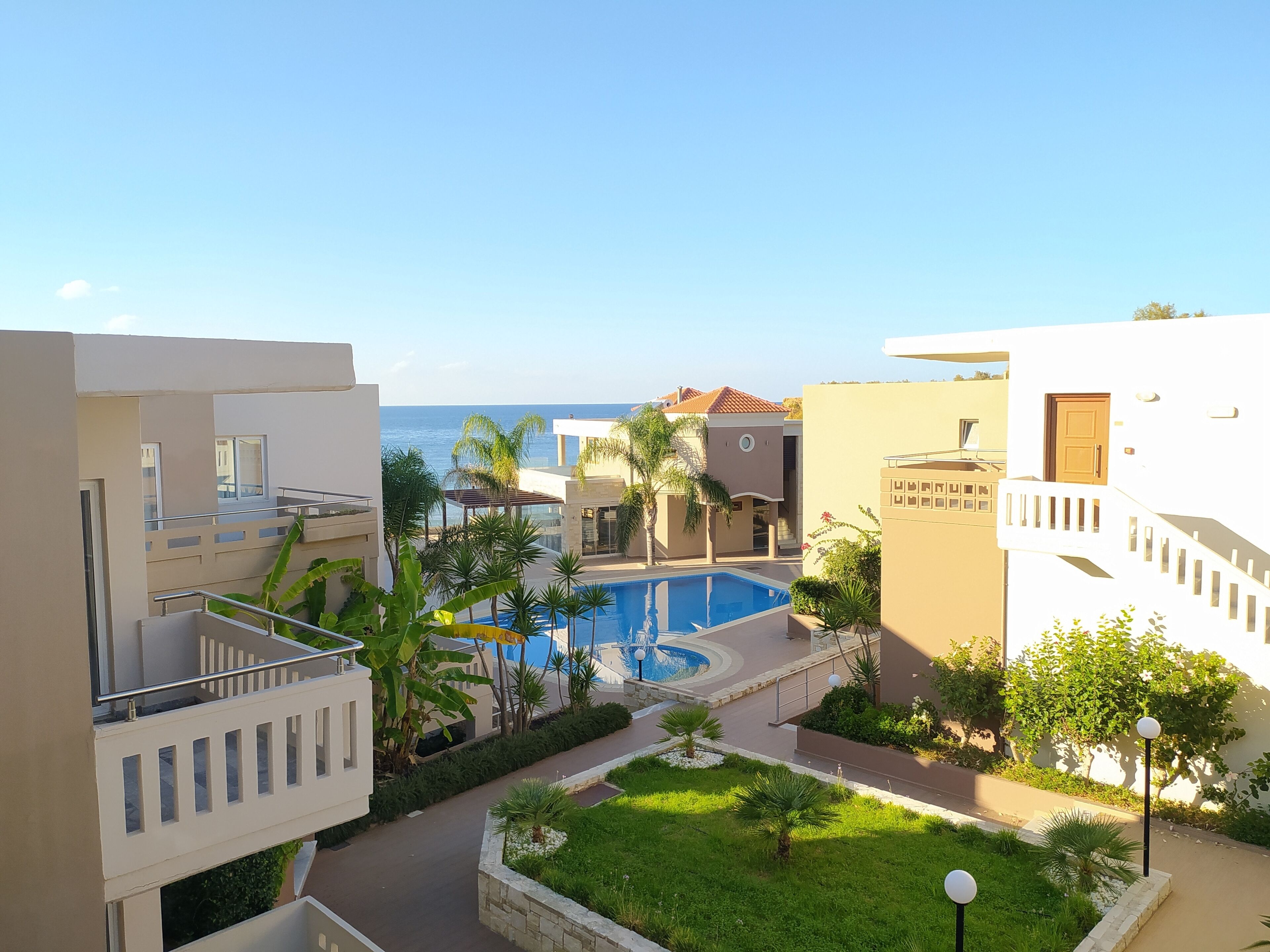 Un complexe hôtelier doté d’une piscine entourée de verdure, offrant une vue dégagée sur la mer et comprenant des bâtiments modernes avec des balcons.