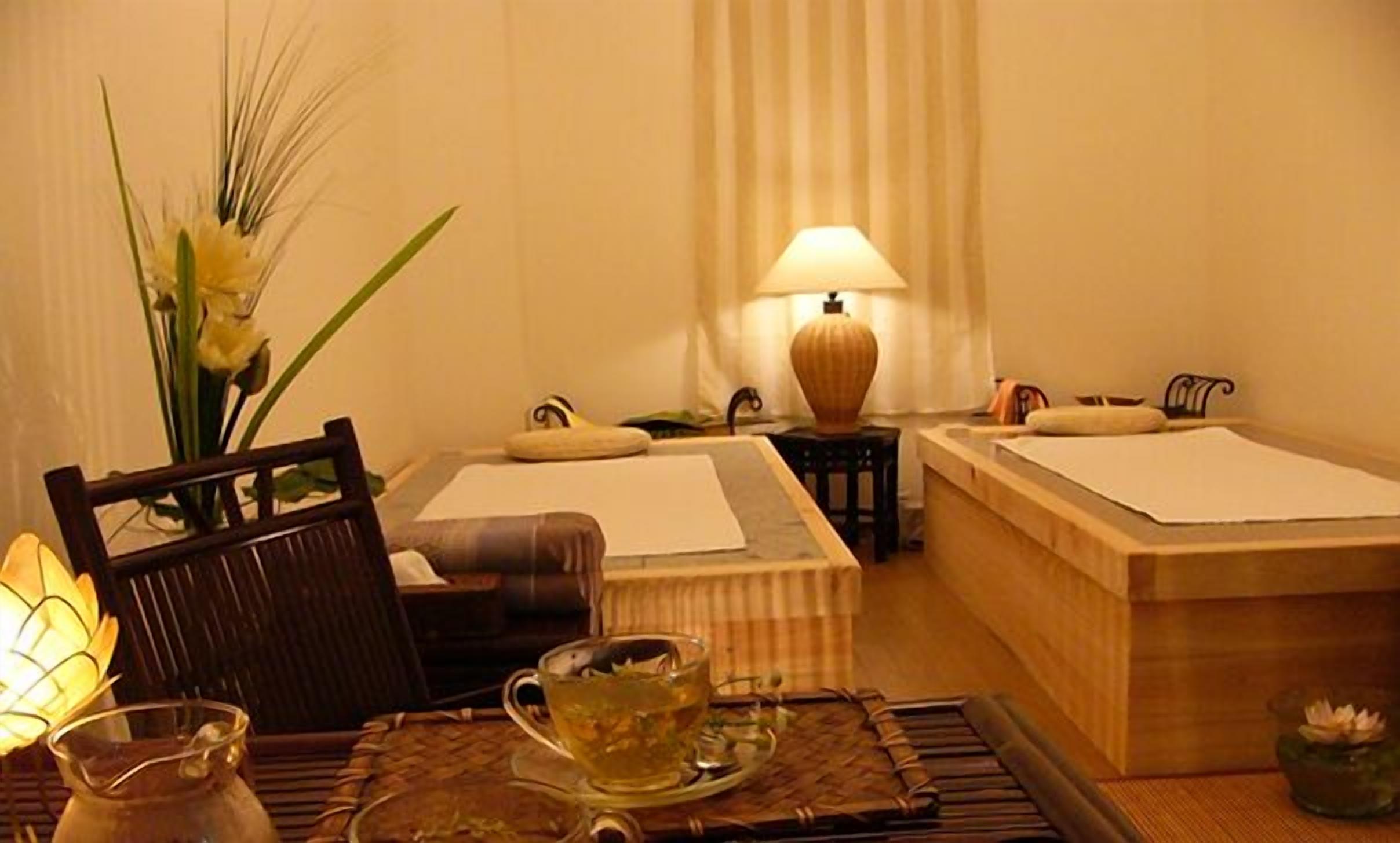 A room with a bed, a chair, a table with a teapot, and a lamp.
