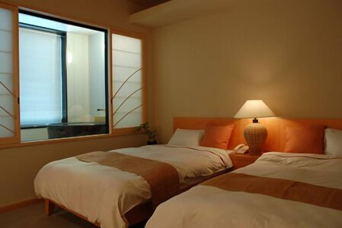 Une chambre d’hôtel avec deux lits, une fenêtre avec des stores, une lampe de chevet et une tête de lit en bois.
