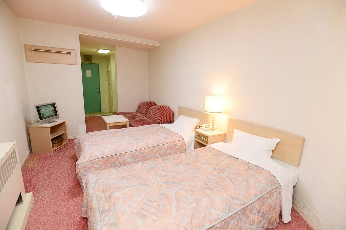 Quarto de hotel com duas camas, uma escrivaninha com computador, uma lâmpada e uma porta verde ao fundo.