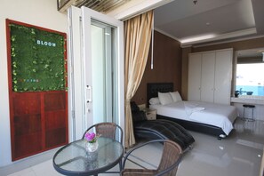 A hotel room with a bed, a glass table with a vase, wicker chairs, a red door with a green wall, and a large window. - Harvia Suites (Jakarta)