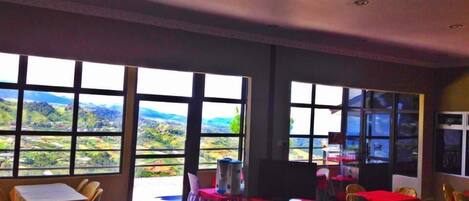 Un comedor con mesas y sillas, ventanas amplias con vistas al campo y un techo con iluminación empotrada.