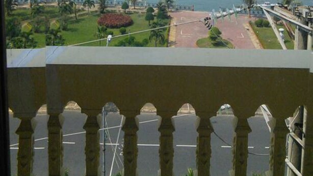 Vista de una zona costera con barcos en el agua, palmeras y una carretera que lleva a un edificio de techo rojo.