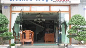 La entrada del Hotel Hoang Ngoc, con fachada de vidrio, muebles de madera en el interior y plantas en macetas en el exterior.