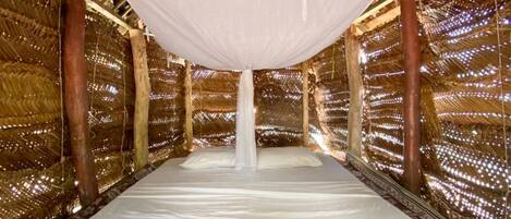 Una habitación con techo de paja, una cama, un dosel blanco colgante y un colorido tapiz tejido.