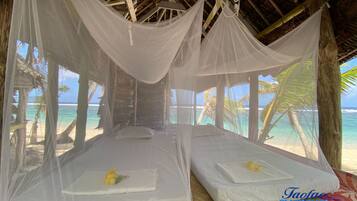 Relax in a serene beachside retreat with two single beds, a thatched roof, and stunning views of the beach and palm trees.