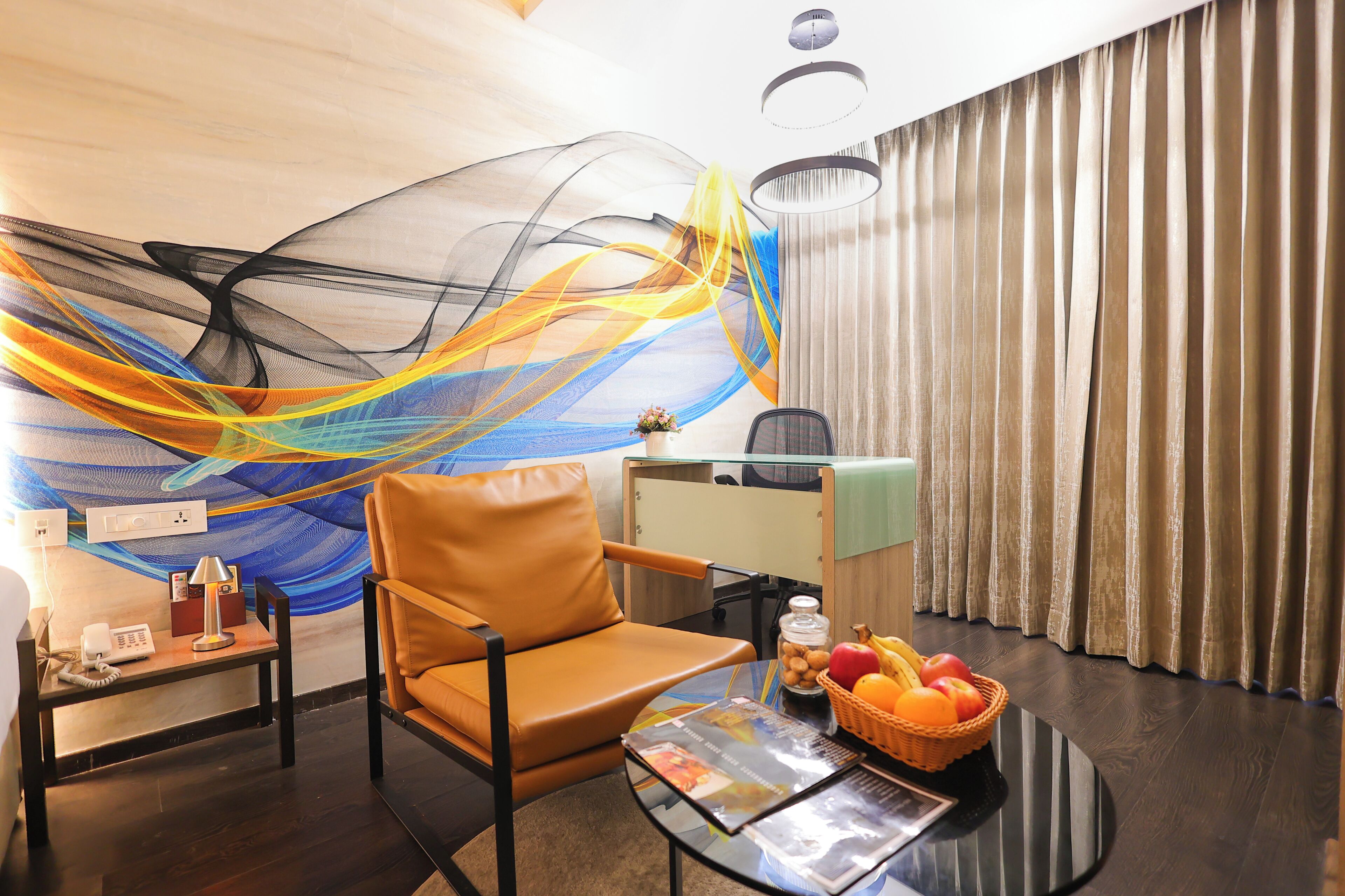 Habitación de hotel con una butaca de cuero, una mesa de centro de vidrio con una revista, una cesta de frutas y una pared con un diseño abstracto y colorido.