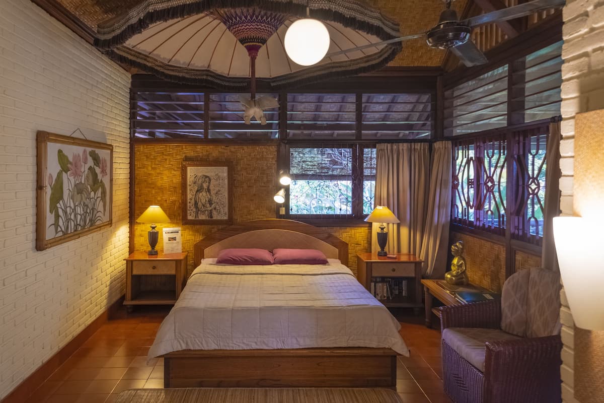 Ein Schlafzimmer mit einem hölzernen Bett, zwei Nachttischen mit Lampen, einem Korbstuhl und einem großen Fenster mit Vorhängen.