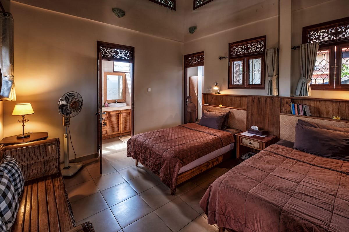 Ein Hotelzimmer mit zwei Betten, einer Sitzecke, einem Ventilator, einer Lampe und einem Fenster mit Vorhängen.