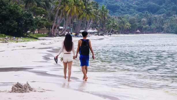 Una pareja caminando por una playa, con palmeras y una cabaña al fondo.