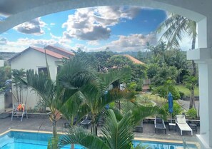 Zona de piscina con sillas de descanso y sombrillas, rodeada de palmeras y vista a una casa y follaje.