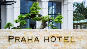Un muro de piedra con el texto "PRAHA HOTEL" frente a un edificio con grandes ventanales y un árbol.