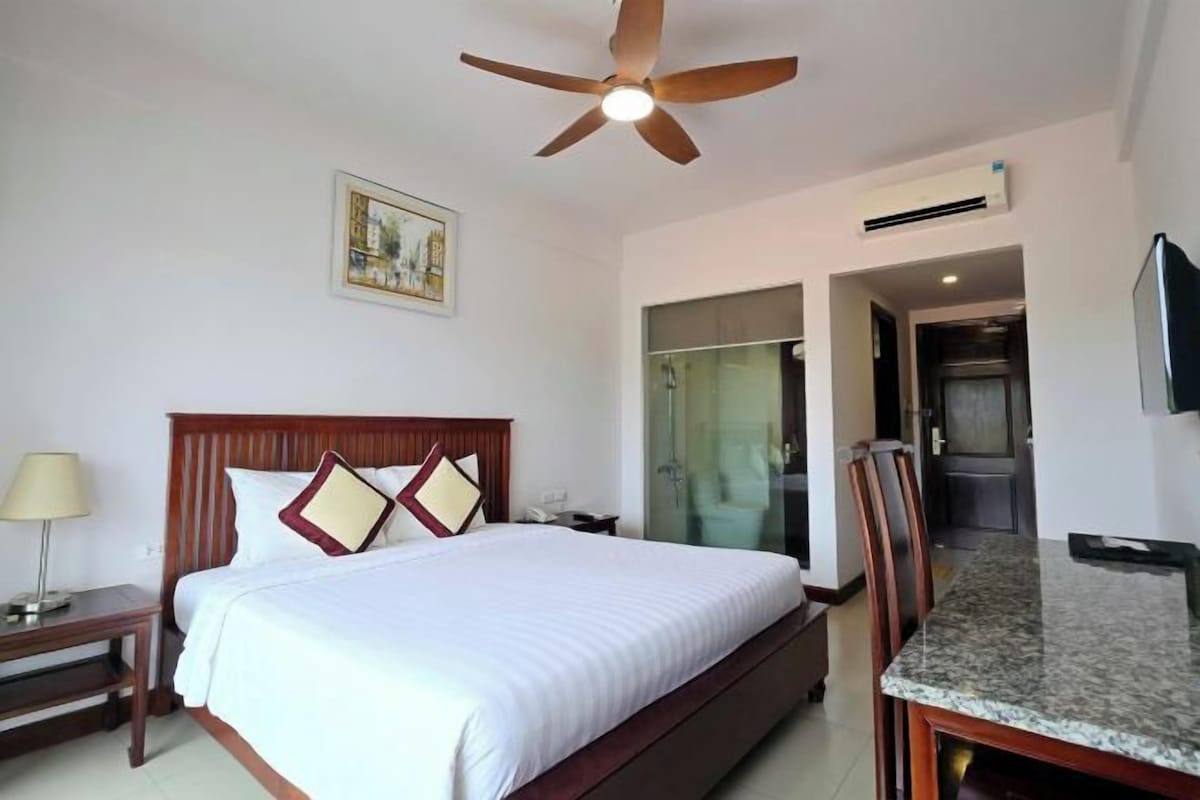 A hotel room with a bed, a desk, a chair, a ceiling fan, an air conditioning unit, a television, and a bathroom with a glass door.