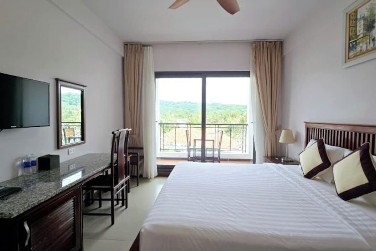 A hotel room with a bed, a desk, a television, and a balcony with a view of greenery.