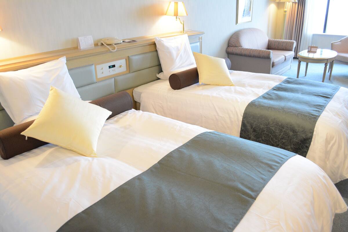 A hotel room with three beds, a bedside table with a lamp, a chair, and a small table.