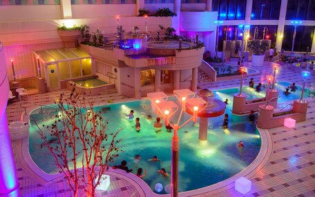 A multi-level indoor pool area with various water features and seating areas, surrounded by balconies and potted plants.