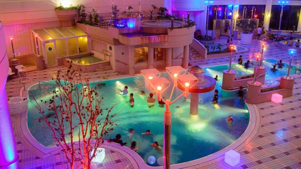 Un área de piscina interior con varios niveles, diversas atracciones acuáticas y zonas de estar, rodeada de balcones y plantas en macetas.