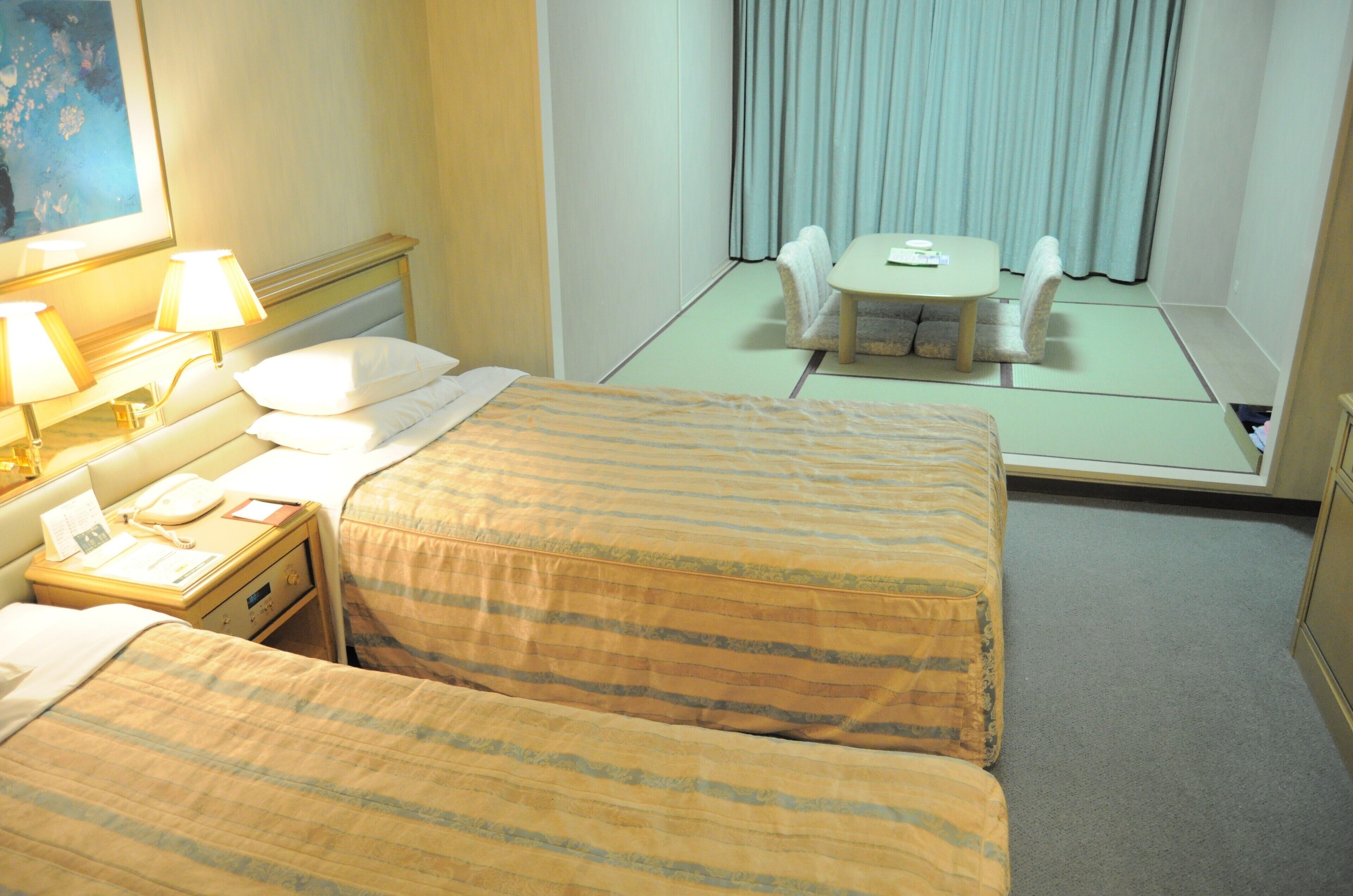 Une chambre d’hôtel avec deux lits, une table de chevet avec une lampe, une petite table avec une chaise, et une vue sur un balcon avec des rideaux.